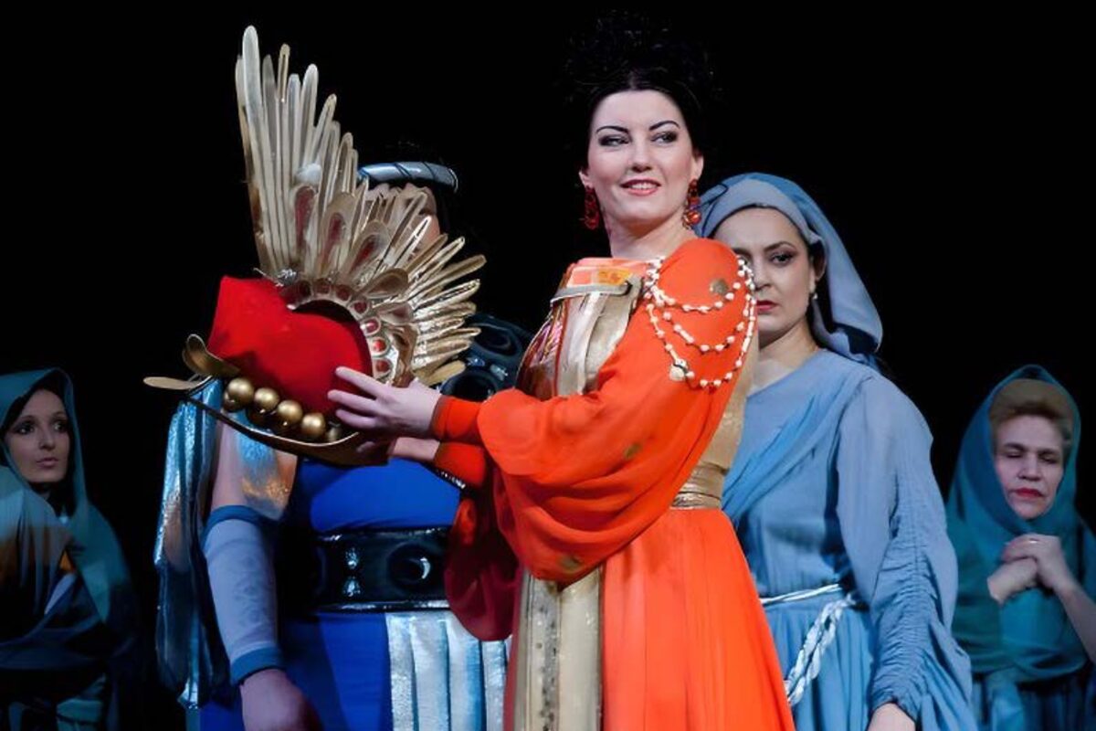 Abigaille, con túnica naranja y corona alada dorada, rodeada por el coro de esclavas hebreas en la producción de Nabucco.
